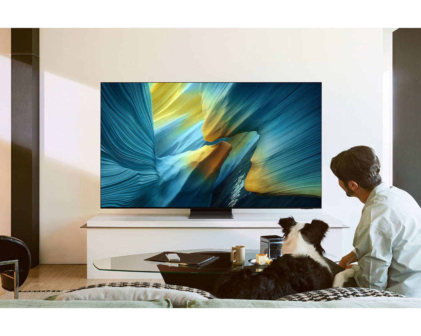 Person and dog sitting on a couch in front of a large TV displaying abstract art.